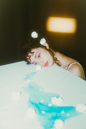 Model laying her head on a white surface with a blue puddle of tears and white roses running down the surface from her eye as she looks away from the camera