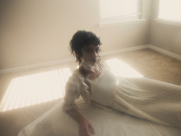 Model is sitting on her side on a small white bed, looking towards the camera. There is a lot of haze in the space, and the bright light source coming from the window adds a dreamy effect