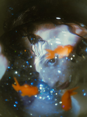 Model looks underwater with goldfish floating on top and her gaze is towards the camera