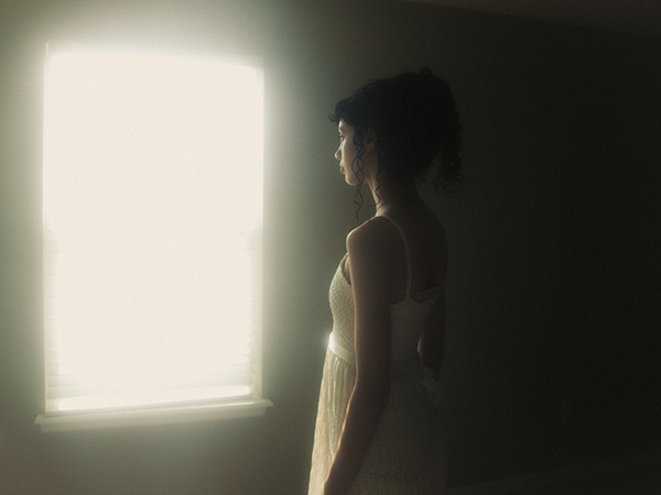 Model is standing straight as she at a bright window, making almost feel like a dreamy portal. There is a lot of haze in the space, and the bright light source coming from the window adds a dreamy effect