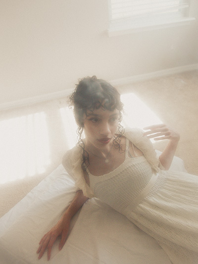 Model is sitting on her side on a small white bed as she gently touches her shoulder, looking away from the camera. There is a lot of haze in the space, and the bright light source coming from the window adds a dreamy effect