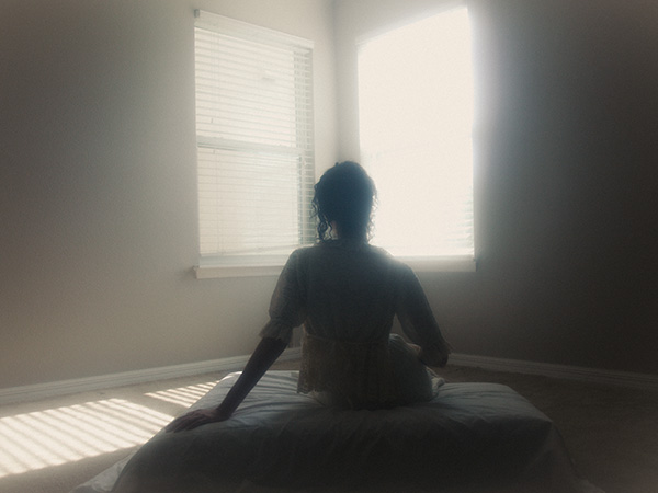 Model is laying on a small white bed facing the camera with her back as she stares at the corner of the room that has two lit up bright windows