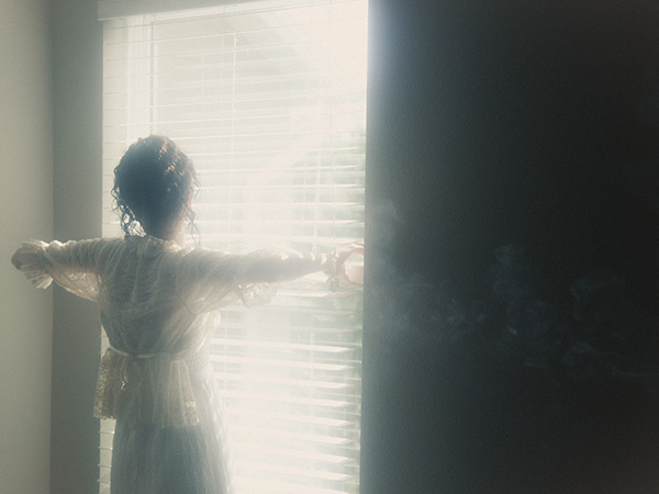 Model is standing as she gently opens the curtains of a window while facing the camera with her back. There is a lot of haze in the space, and the bright light source coming from the window adds a dreamy effect