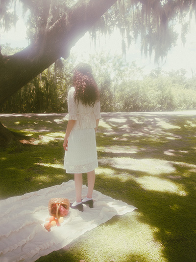 Model is standing on her picnic sheet facing the camera with back. There is a fruit picnic basket by her feet with fruit organically pouring down from it on