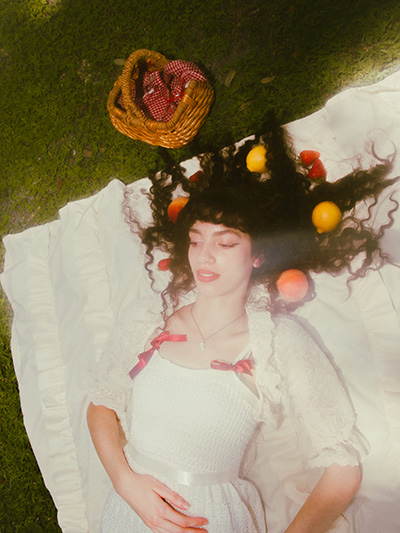 Model is laying down on her picnic sheet gazing away from the camera. She has peaches, lemons, and strawberries surrounding her curly hair and a picnic basket is nearby